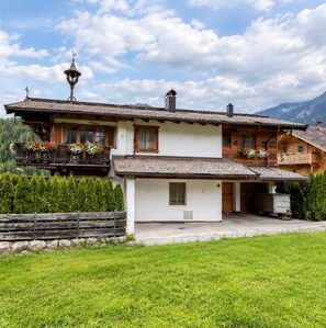 A two-story chalet with a balcony, a chimney, and a mountain backdrop.