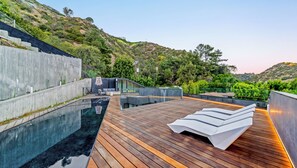 Piscina infinita en la azotea con cubierta de madera y sillas de descanso, rodeada de vegetación y una ladera.