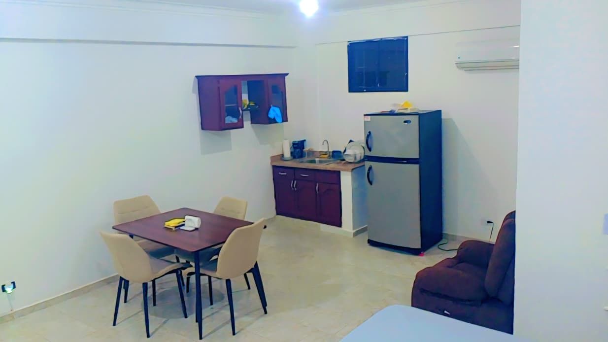 Een knus woongedeelte met een eettafel, stoelen, een kitchenette met een koelkast, en een zithoekje.