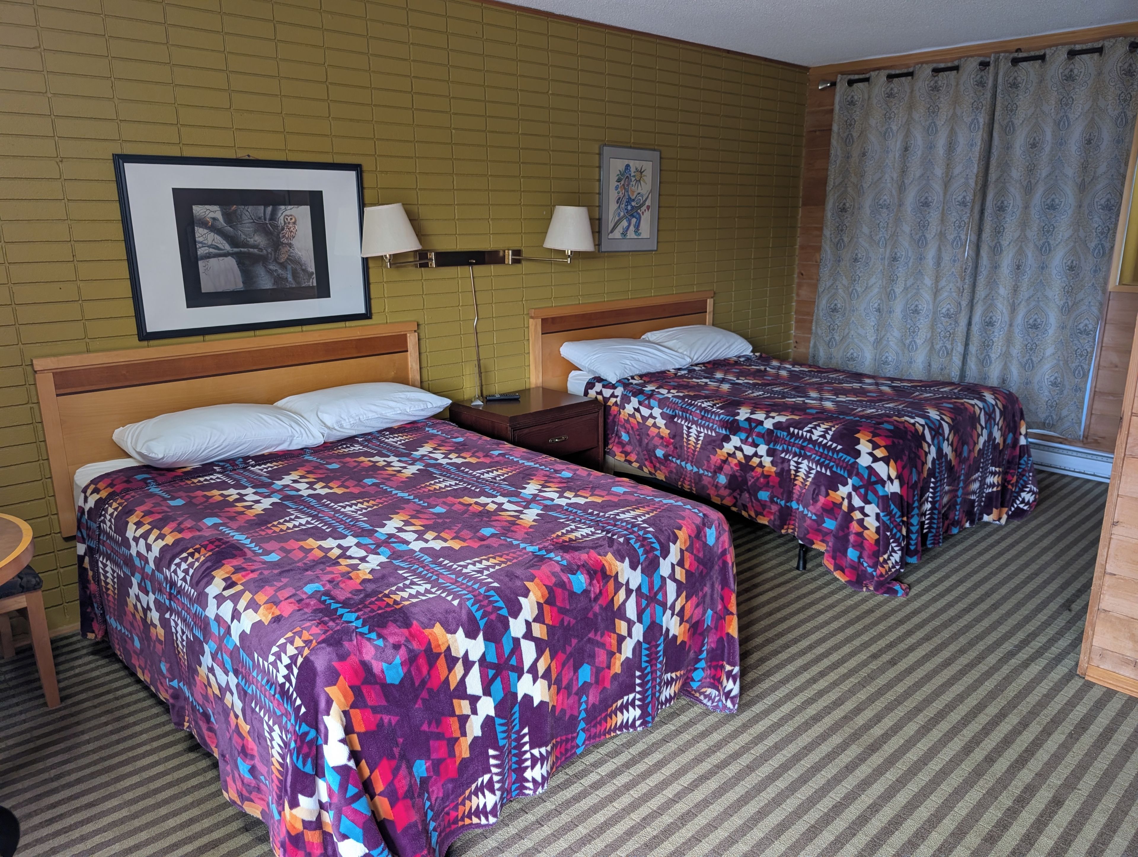A hotel room with two beds, a wooden headboard, a nightstand, a framed picture, and a patterned curtain.