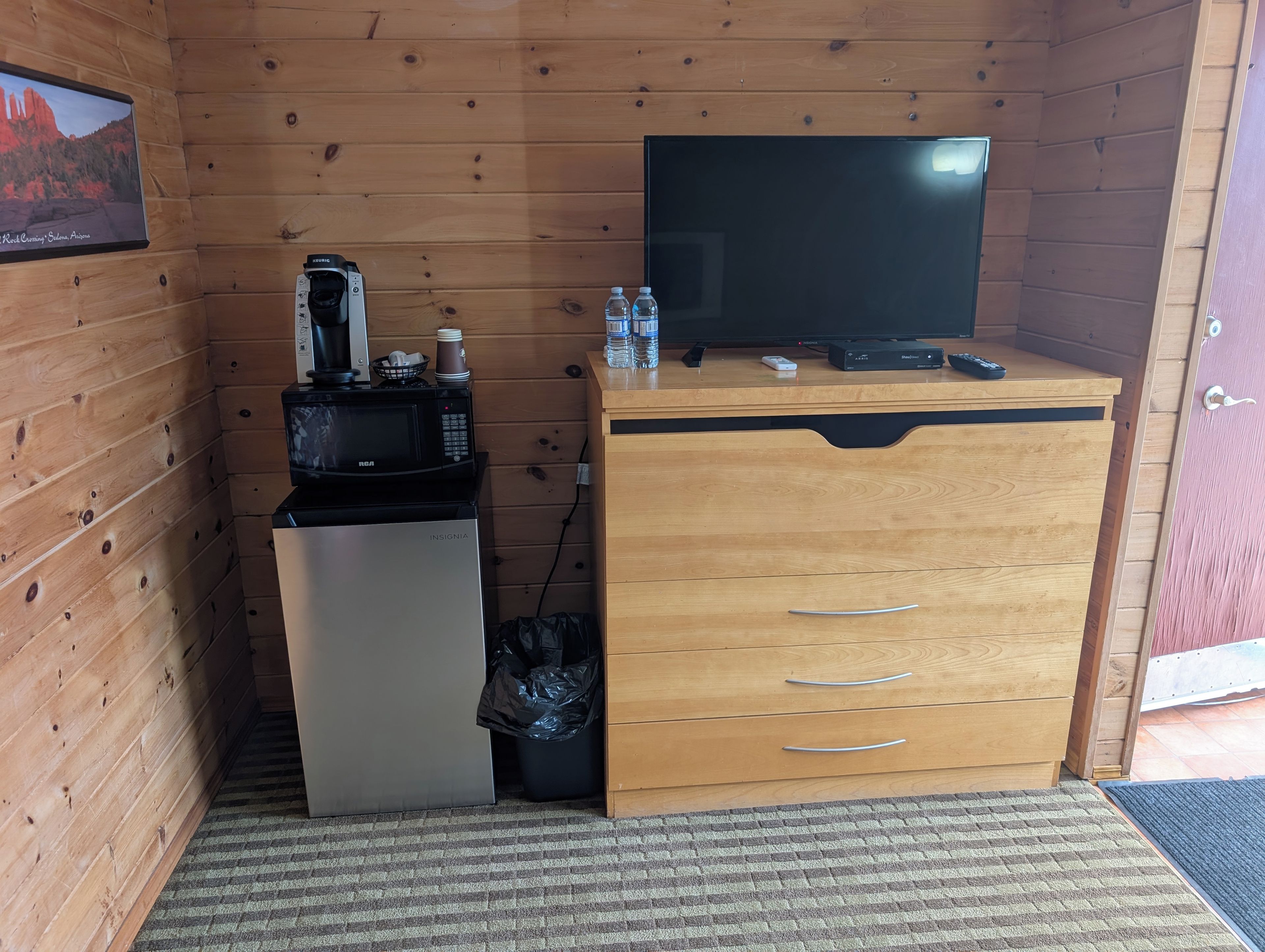 A compact kitchenette with a microwave, coffee maker, and a small refrigerator. A television is mounted on the wall, and there are wooden cabinets with drawers.