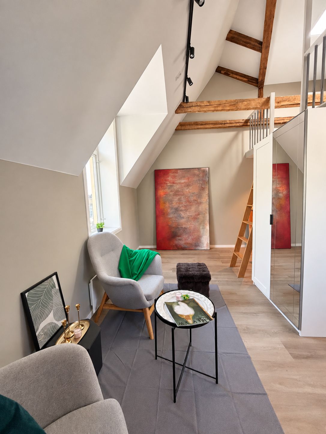 A modern loft with a sloped ceiling, a grey armchair with a green throw, a small round table with a book, and a ladder leading to an upper level.