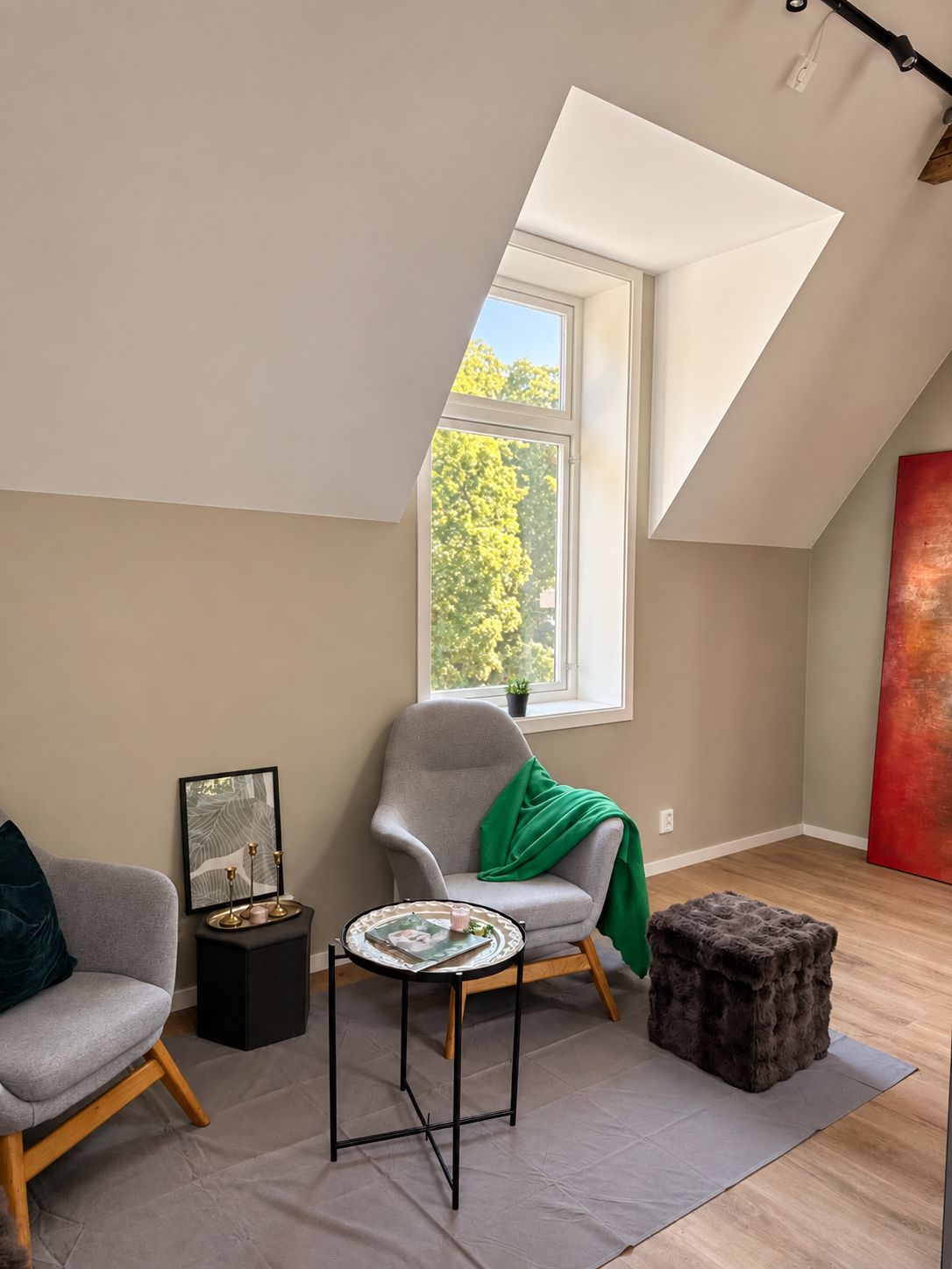 A modern loft bedroom with a sloped ceiling, a large window with a view of trees, a grey armchair, a small round table, and a red abstract painting.
