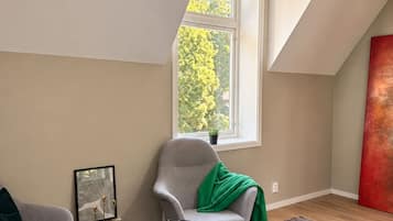 A modern loft bedroom with a sloped ceiling, a large window with a view of trees, a grey armchair, a small round table, and a red abstract painting.
