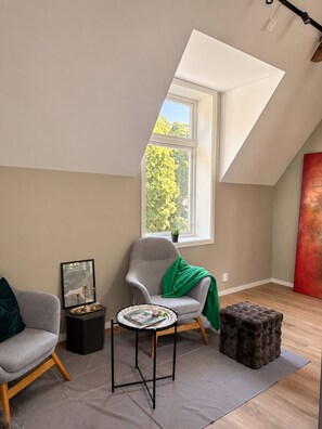 A modern loft bedroom with a sloped ceiling, a large window with a view of trees, a grey armchair, a small round table, and a red abstract painting. - Modern Private Studio with Loft Central Oslo (Oslo)