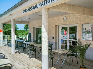 Un bar-restaurant en plein air, avec des tables et des chaises, une horloge et une vue sur la verdure.