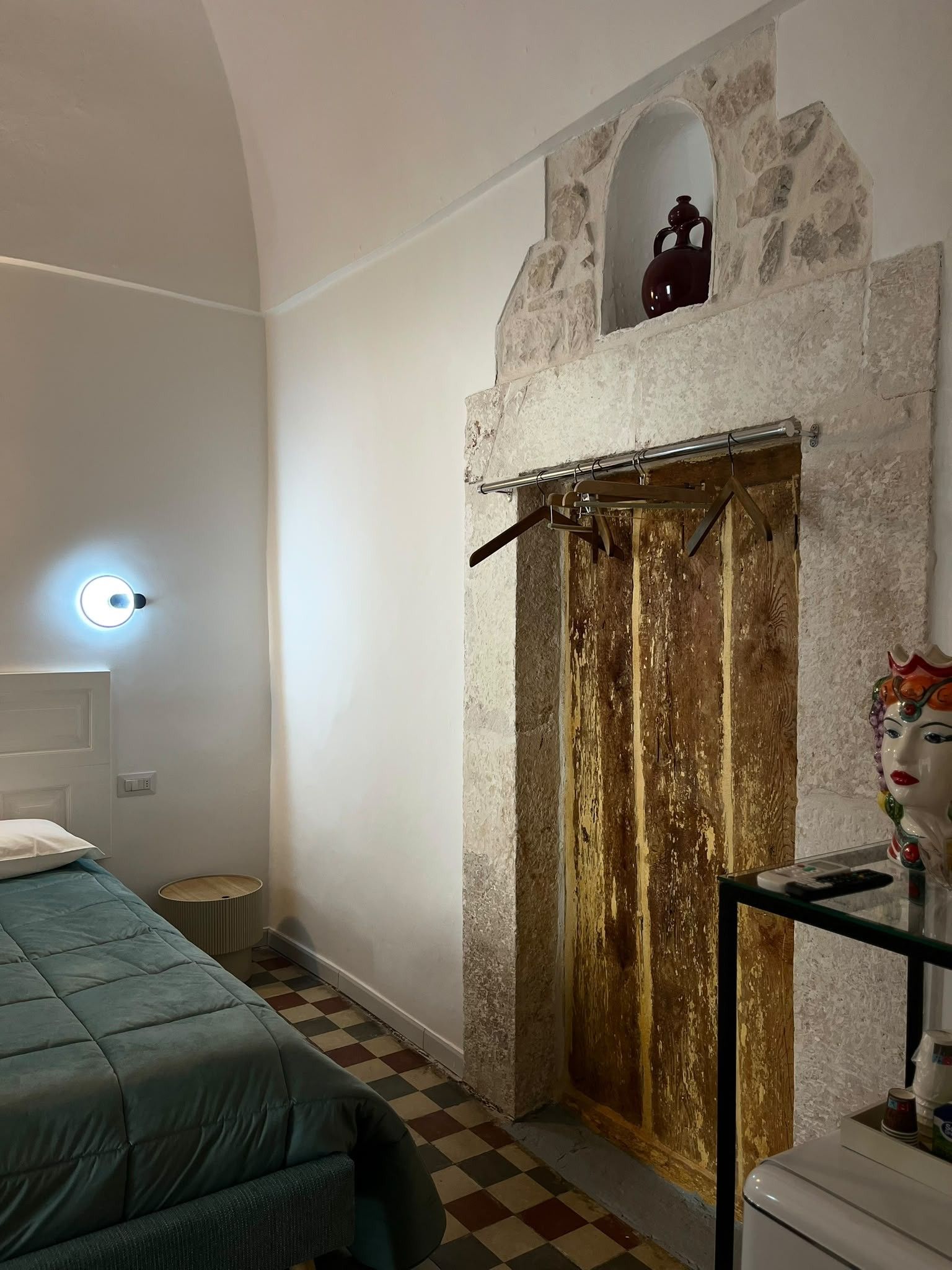 A room with a bed, a stone wall, a wooden door, and a small window.