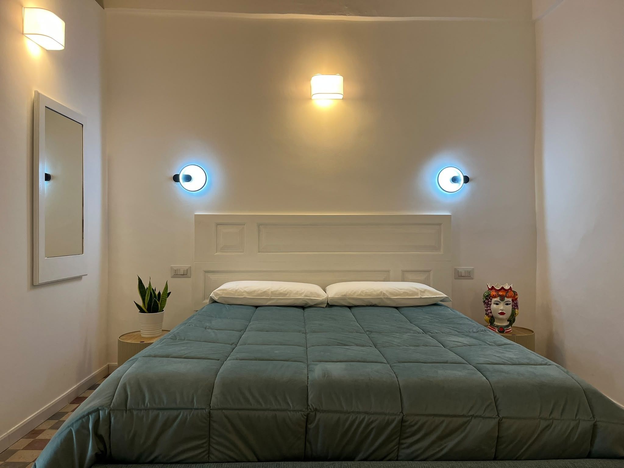 A bedroom with a bed, two wall-mounted lights, a nightstand with a plant, and a decorative item.