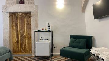 A room with a checkered floor, a green sofa, a small fridge, a coat rack, and a stone wall with a wooden door.
