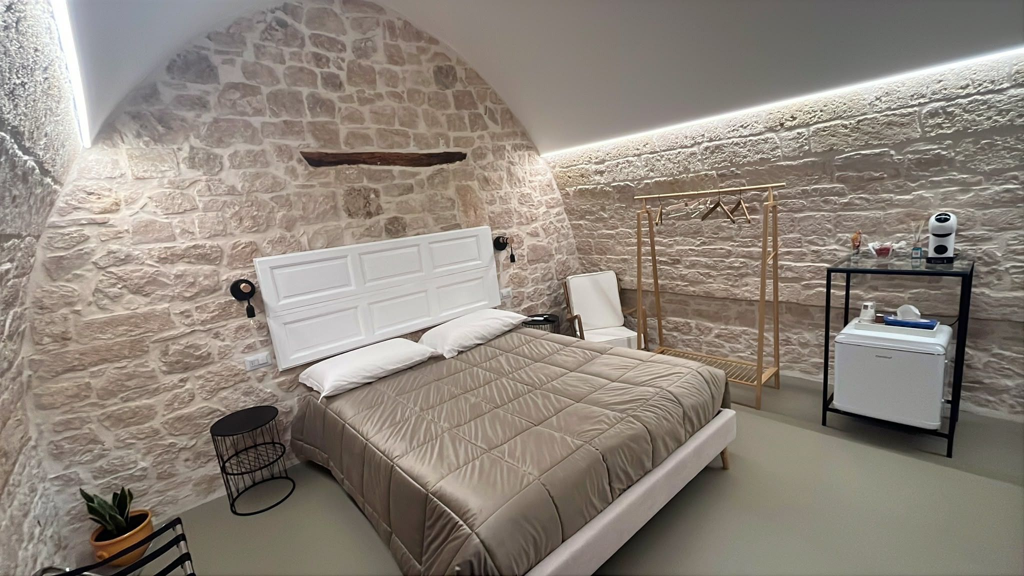 A bedroom with a stone wall, a bed with a white headboard, a bedside table, a chair, a small table with a lamp, and a wardrobe.
