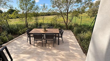 En udendørs terrasse med et rektangulært bord og stole, omgivet af en have med træer og et hegn.