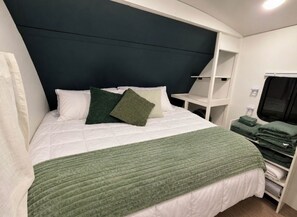 Un lit bien fait, avec une couverture verte et des oreillers, une tête de lit blanche et une table de chevet blanche sur laquelle est posée une serviette verte pliée.