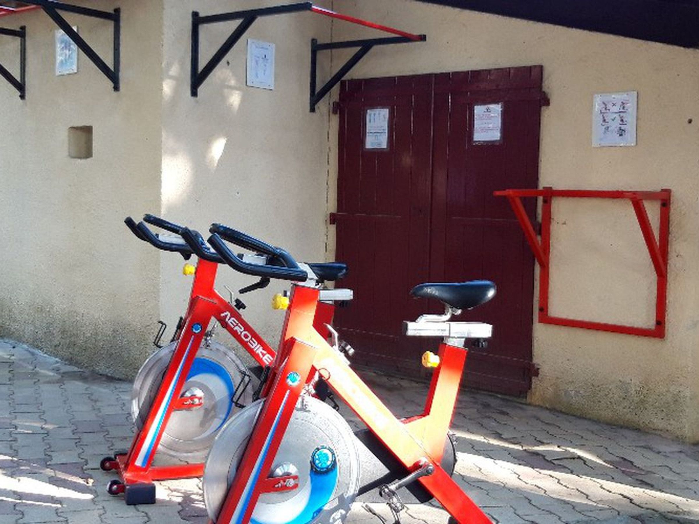 A stationary exercise bike with a red frame and blue accents, parked outside a building with a red door and a small window.