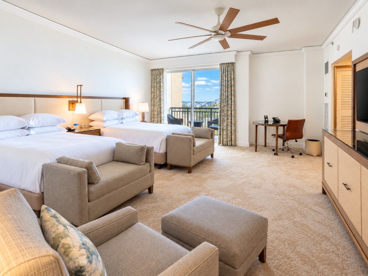 A hotel room with two beds, a desk, a television, and a balcony with curtains.