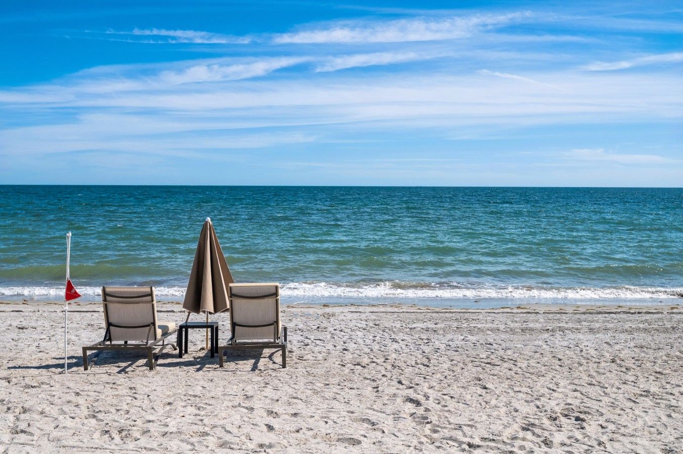 A beach with two chairs and a table, a beach umbrella, and a flag on the sand.