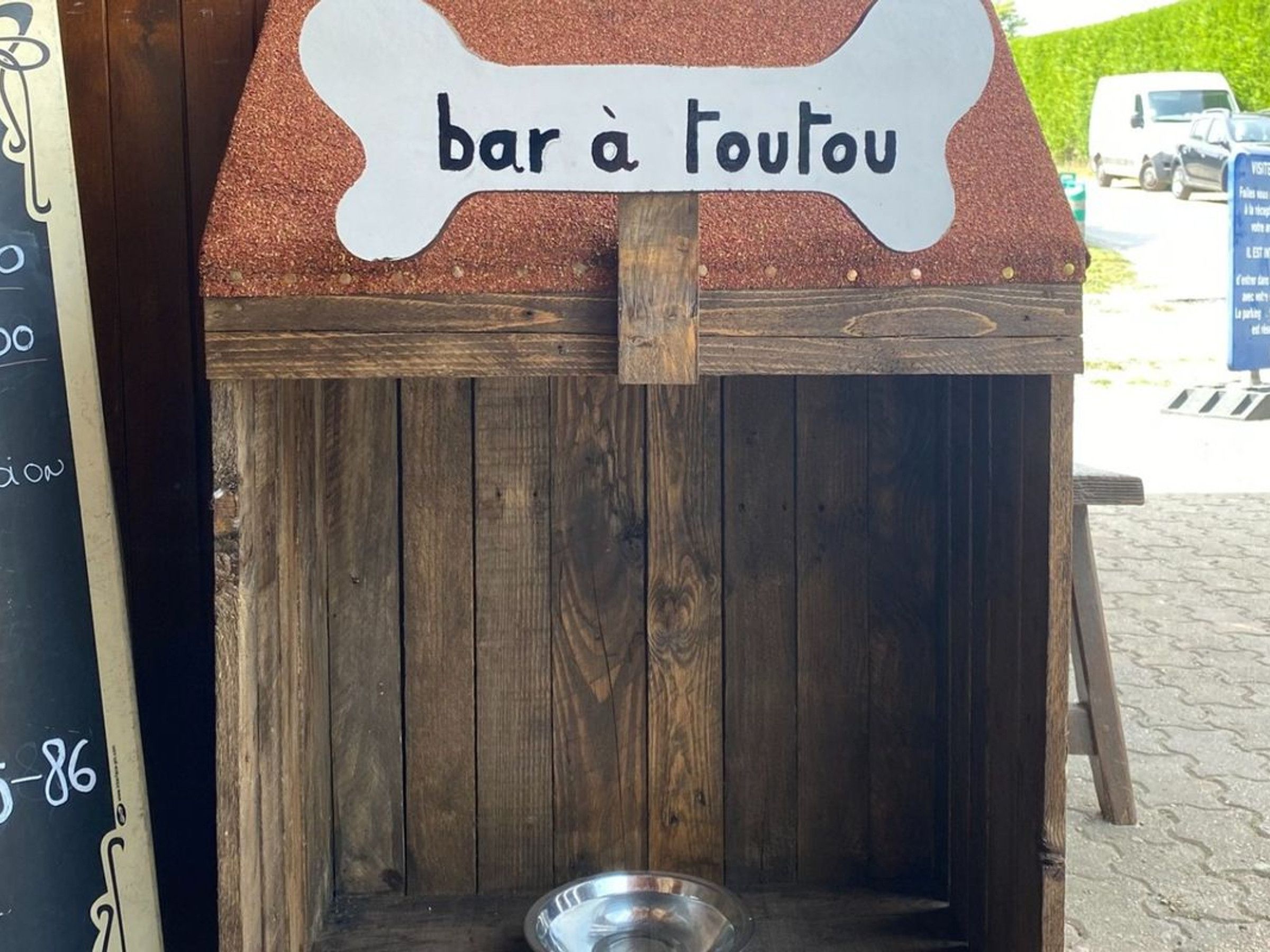 Une structure rustique en bois, sur laquelle est accrochée une enseigne indiquant « bar à chiens », et un bol en métal posé sur le sol.