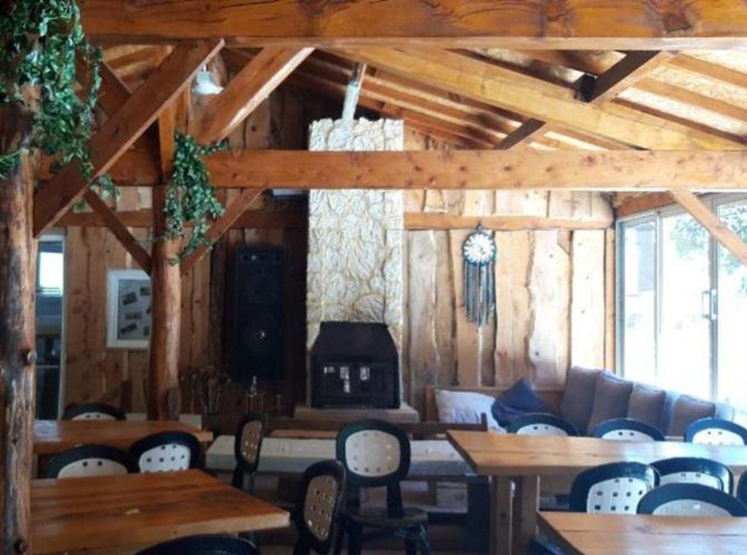 A rustic wooden interior with exposed beams, a stone wall, and a fireplace. Tables and chairs are arranged for dining.