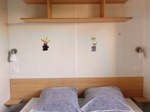 A bedroom with a wooden headboard, two bedside tables, a bed with gray bedding, and wall-mounted light fixtures. - Mobile home with air conditioning - 2 guests (Angicourt)