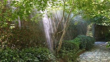 A stone pathway leading through a lush garden with dense foliage and a waterfall.