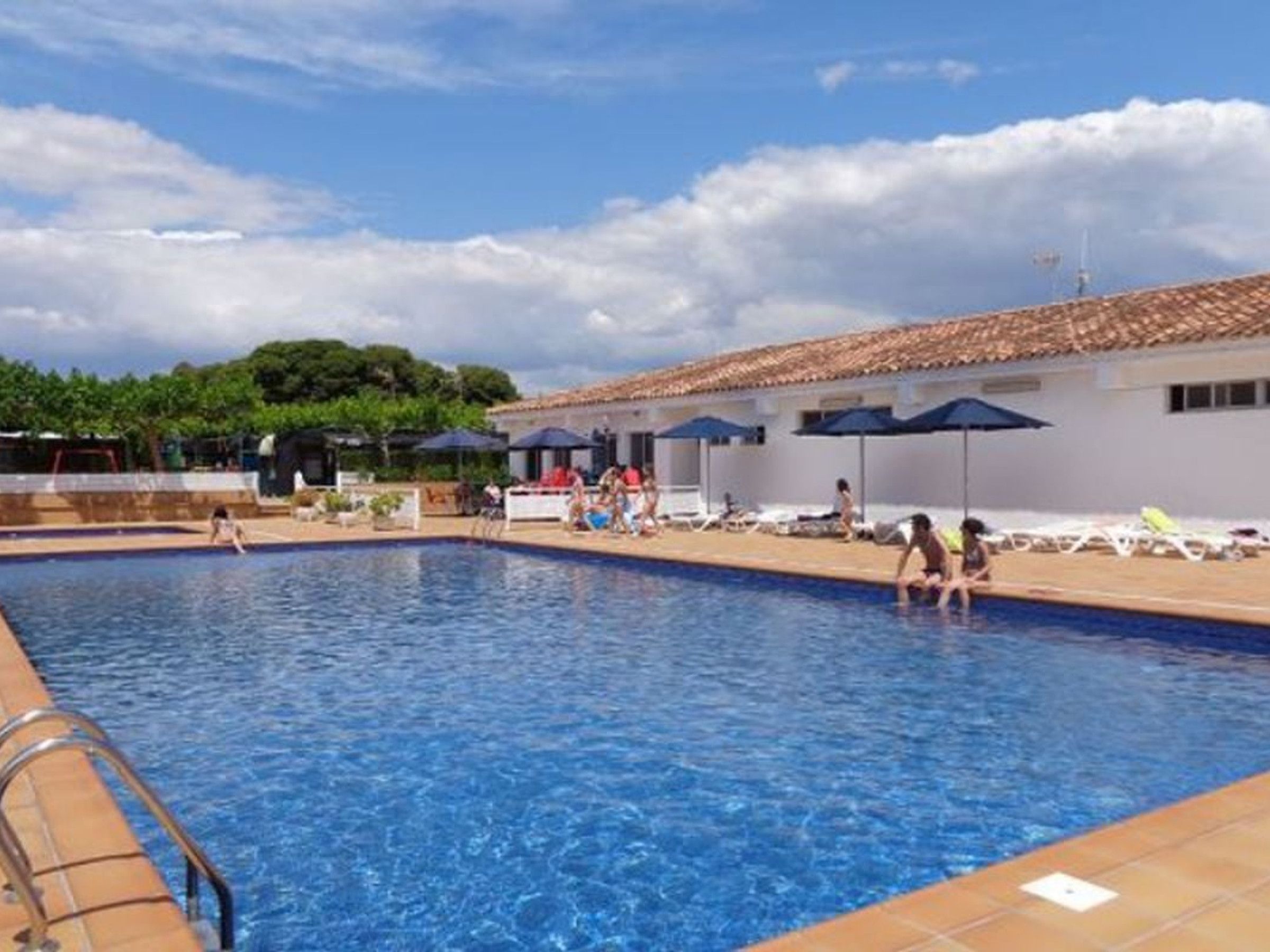Zona de piscina con suelo de baldosas, sillas de descanso y sombrillas.