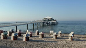 A beach with beach huts and a pier leading to a modern building with a dome roof.