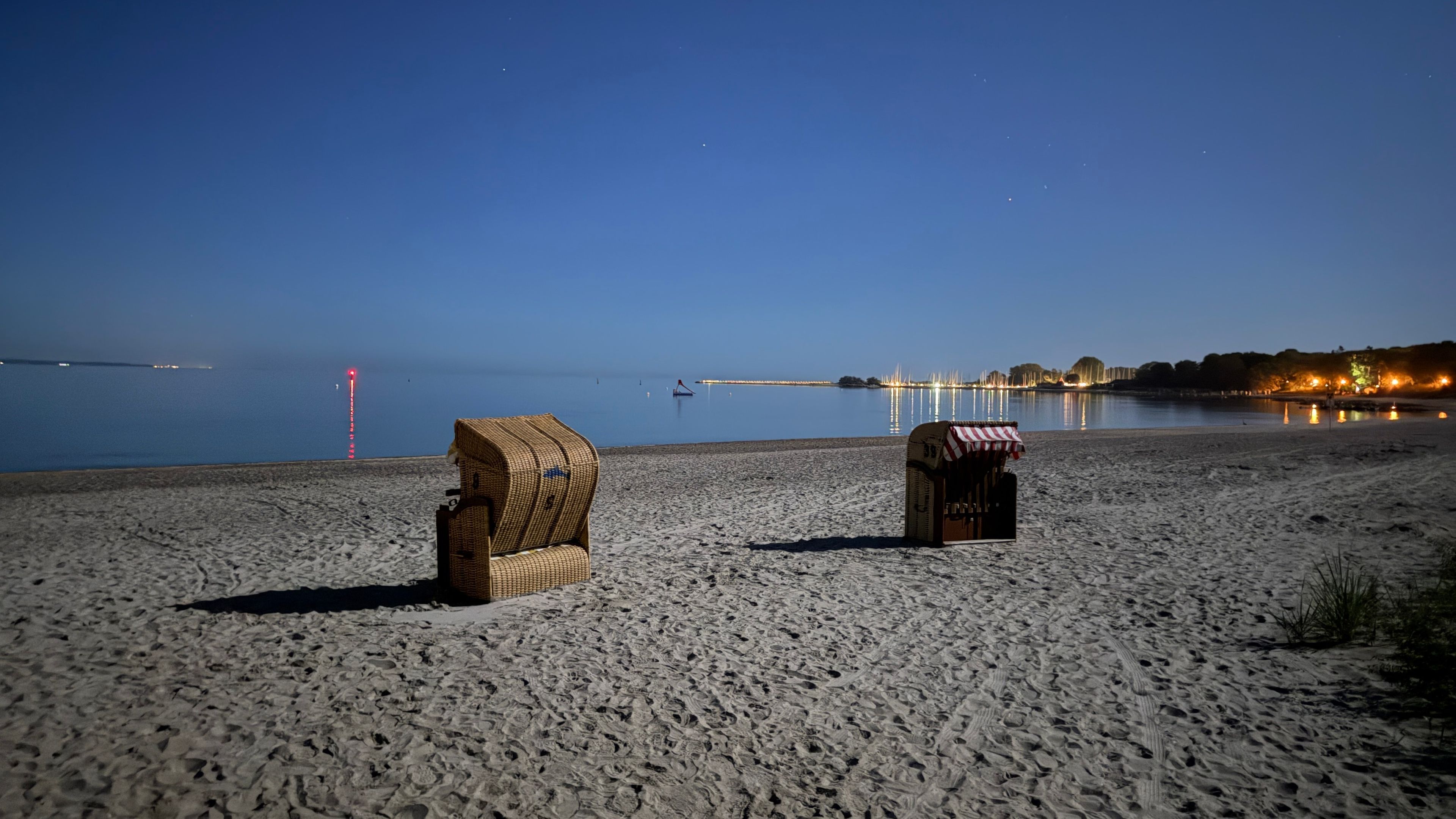 A beach with two beach chairs and a small structure, likely a snack or drink stand, with a calm sea and distant lights in the background.