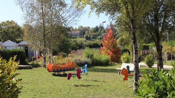 Un'area verde con alberi, un'area giochi con scivolo e altalene, e un edificio sullo sfondo.