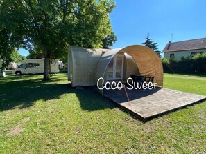 Eine kleine, halbkreisförmige Holzhütte mit einer flachen Plattform davor, vermutlich ein Zelt oder eine kleine Hütte, eingebettet in eine grüne Wiese mit Bäumen und einem Haus im Hintergrund.