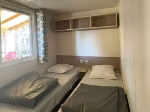 A small room with two single beds, a window with a curtain, and a built-in shelf above the beds. - Spacious mobile home with terrace - 8 guests (Athée)