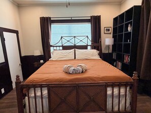 Una cama bien hecha con colcha marrón y almohadas, cabecera de madera, mesita de noche con lámpara y estantería con libros.