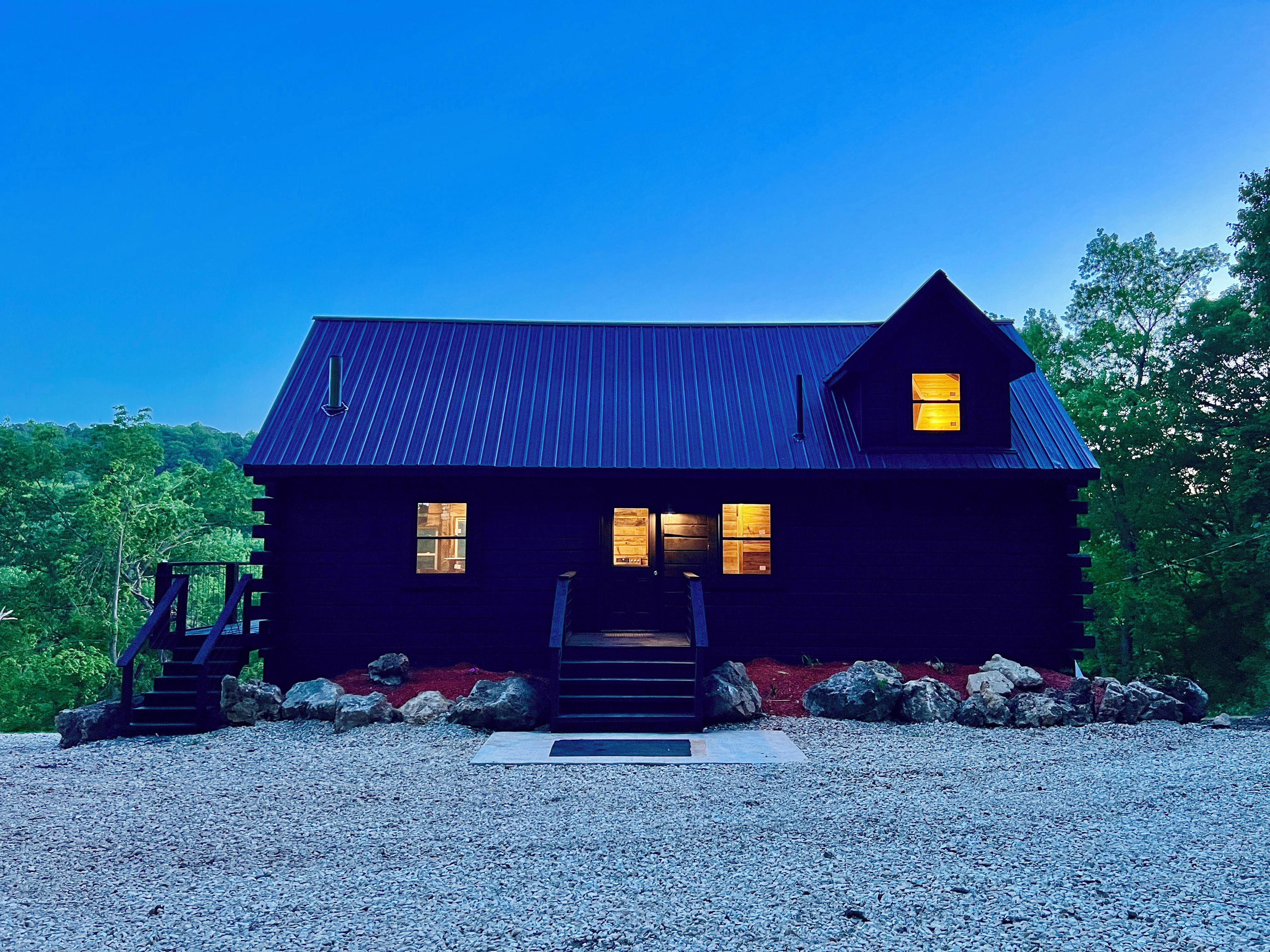 A dark-colored cabin with a metal roof, a front porch, and a gravel driveway.