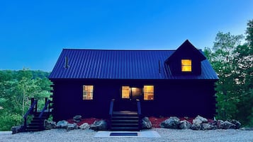 A dark-colored cabin with a metal roof, a front porch, and a gravel driveway.