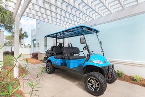 Buggy de golf blau estacionat en una zona coberta, amb un edifici blanc al fons.