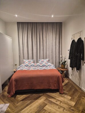 Ein Schlafzimmer mit einem Bett, Vorhängen, einem Nachttisch, einer Lampe, einem Topfpflanze und einem Kleiderständer.