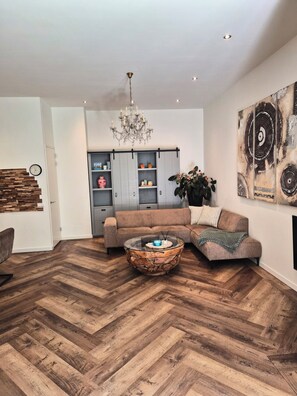 Una sala de estar moderna con piso de madera en patrón chevron, una mesa de centro de vidrio, un sofá marrón y una araña.