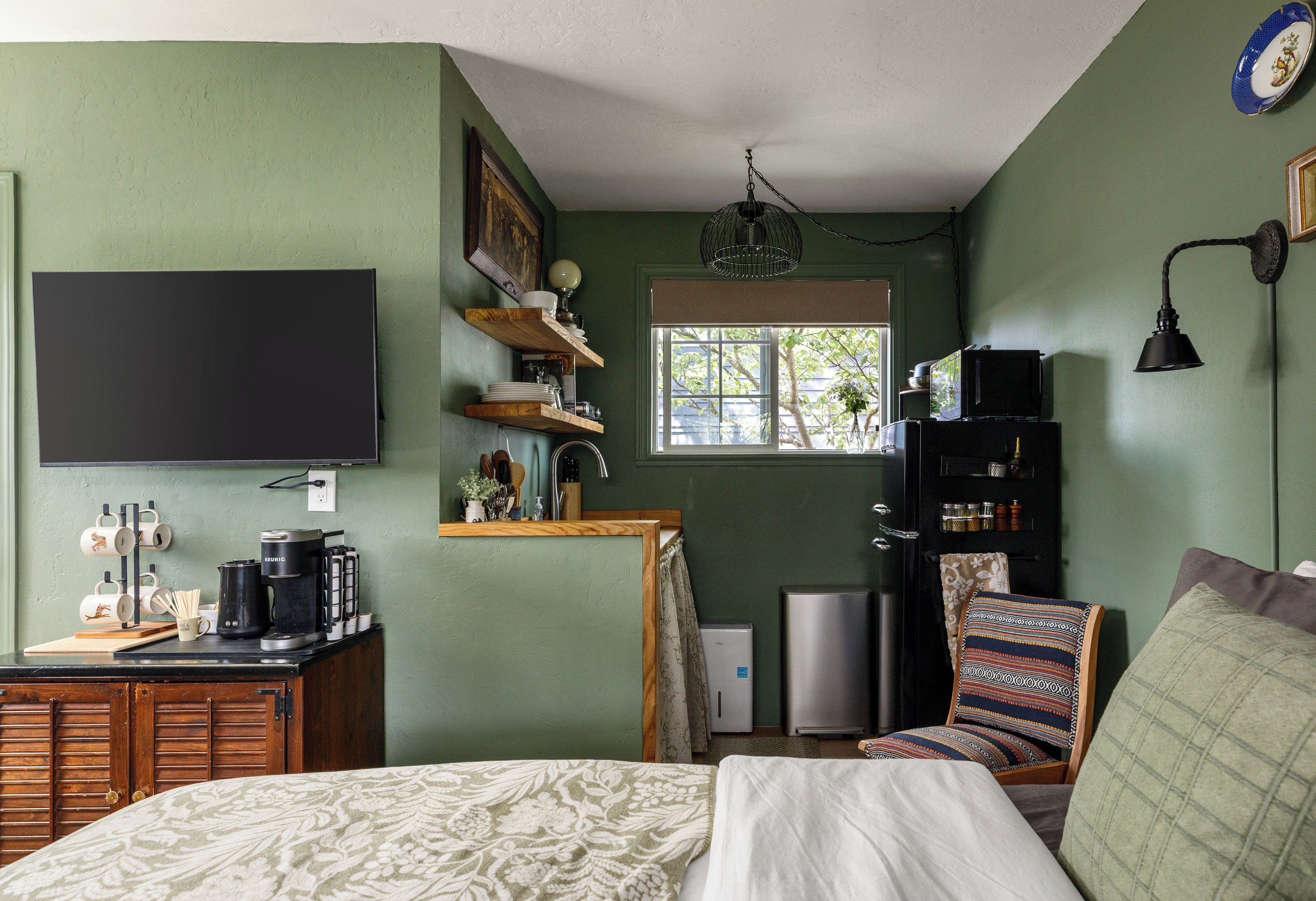 Un dormitorio con cama, televisor montado en la pared, ventana con vista a árboles y una zona de kitchenette.