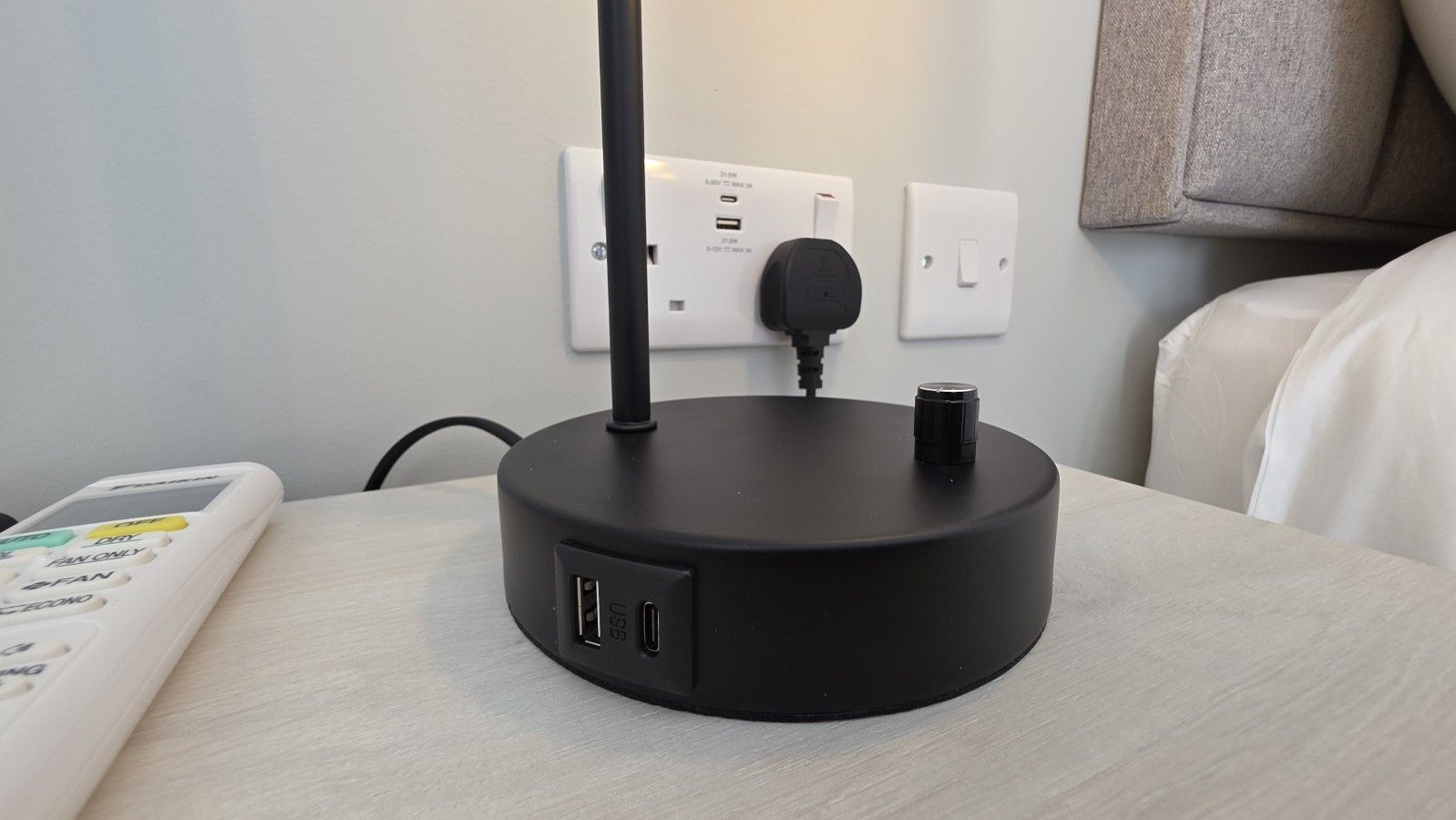 A black portable Wi-Fi router on a wooden table with a remote control beside it, a power outlet, and a light switch on the wall.