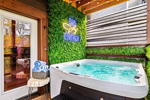 Un bain à remous avec un panneau néon sur lequel on peut lire « BIG POLLY ENERGY », le tout se détachant sur un fond de terrasse en bois et de verdure.