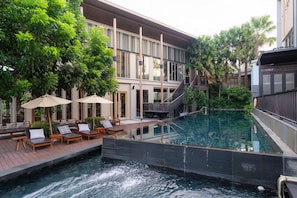Un área de piscina en la azotea con sillones de descanso y sombrillas, rodeada de exuberante vegetación y un edificio moderno.