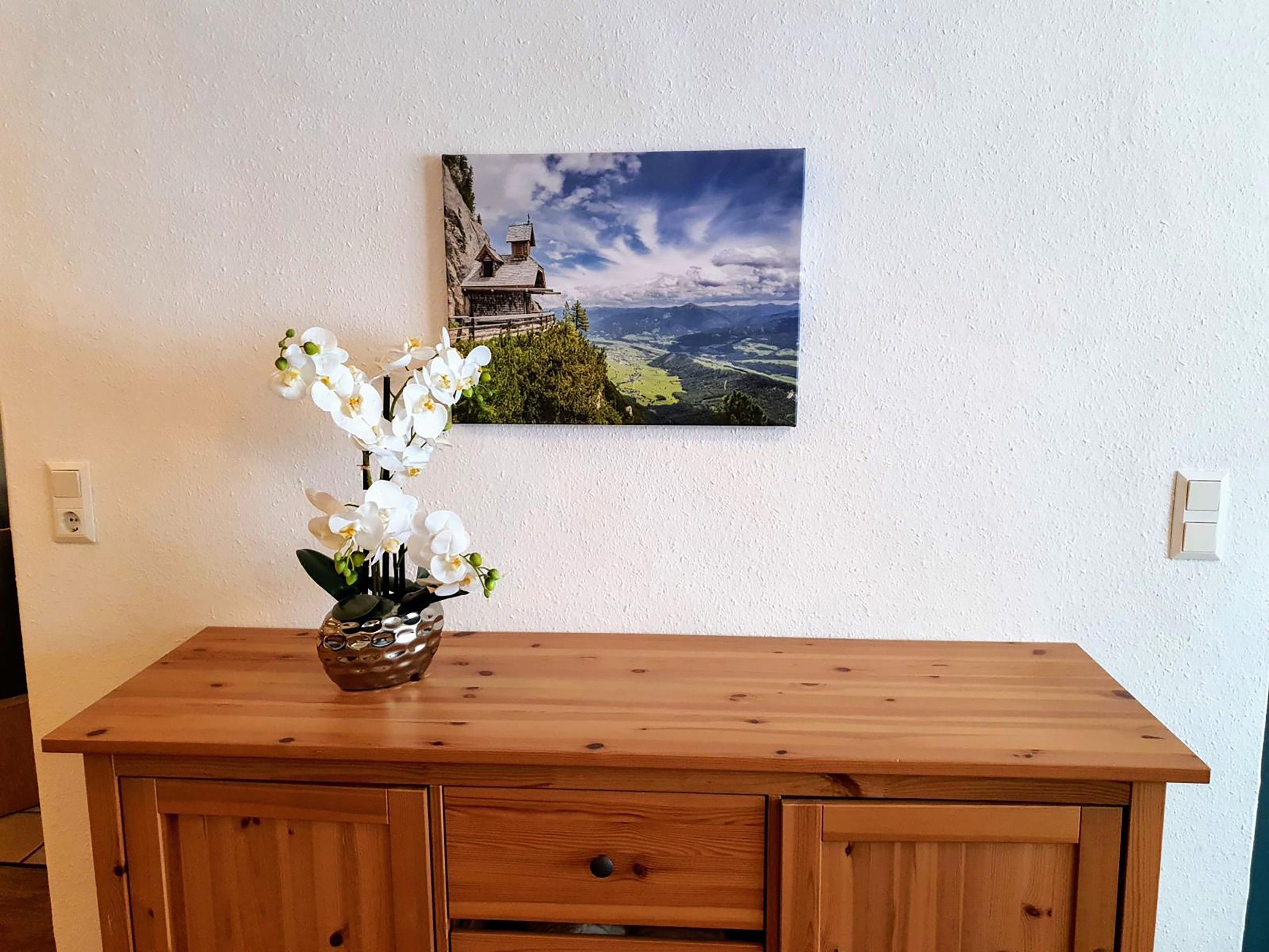 Eine Holzkommode mit einer Vasen voller weißer Orchideen und einem Landschaftsbild an der Wand.