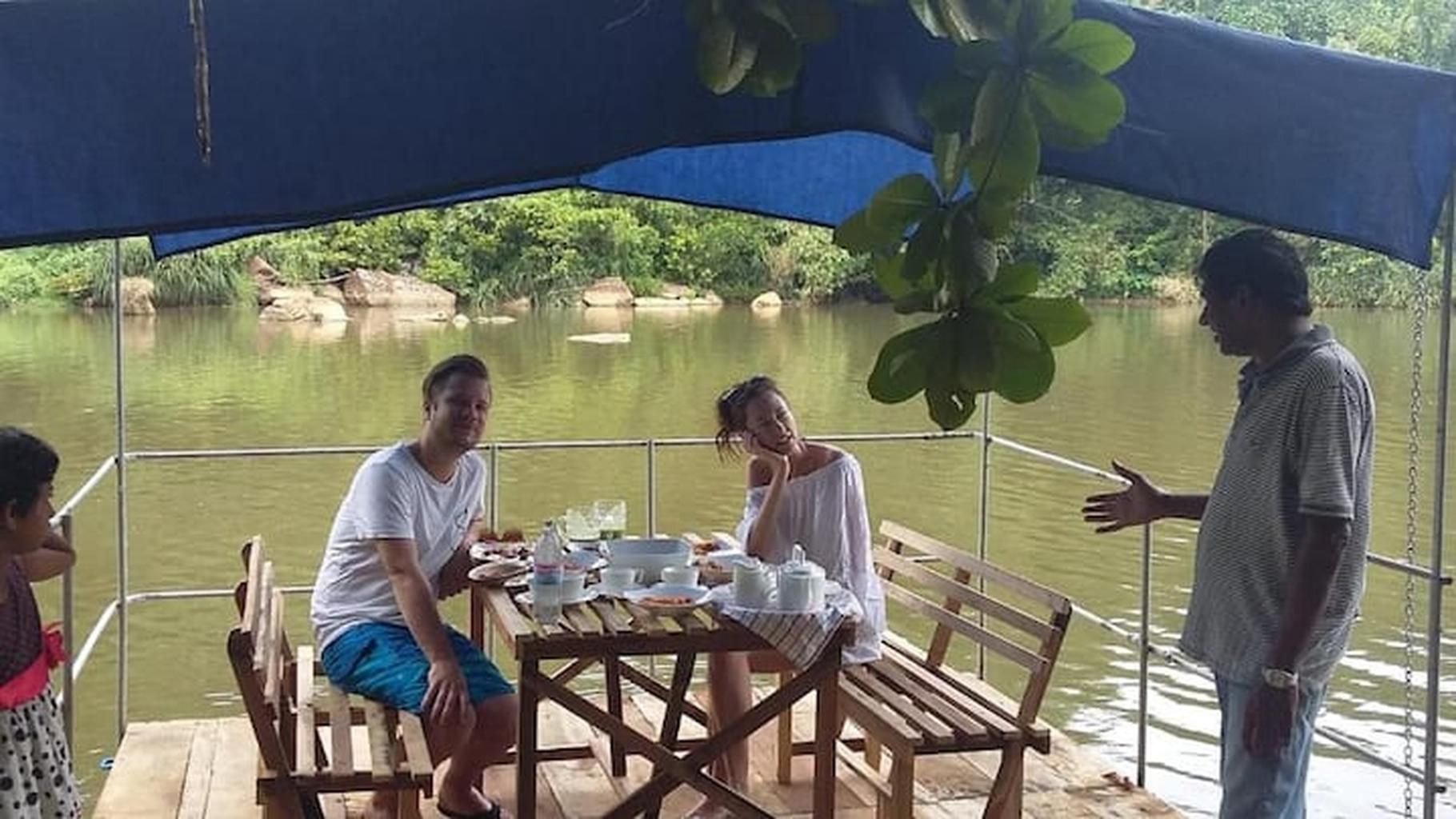 Un groupe de personnes est réuni autour d’une table dressée pour deux sur un bateau, entouré d’une étendue d’eau calme et de verdure.