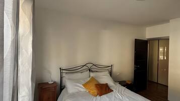 Quarto com cama, mesa de cabeceira, piso de madeira e porta que dá para outro cômodo.