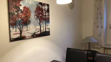 A dining area with a table, chairs, a wall-mounted painting, a wall clock, and a table lamp.