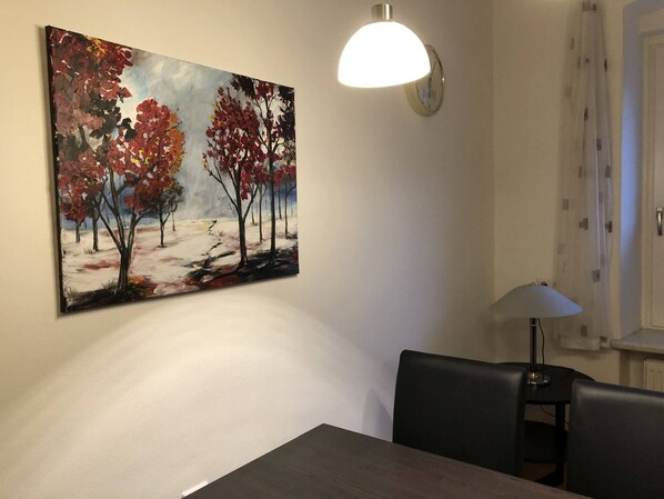 A dining area with a table, chairs, a wall-mounted painting, a wall clock, and a table lamp. - Apartment zur Schmiede (Gröbming)