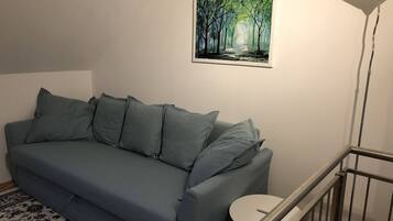 A grey sofa with multiple cushions, a small round table, a painting of a forest, and a modern floor lamp.