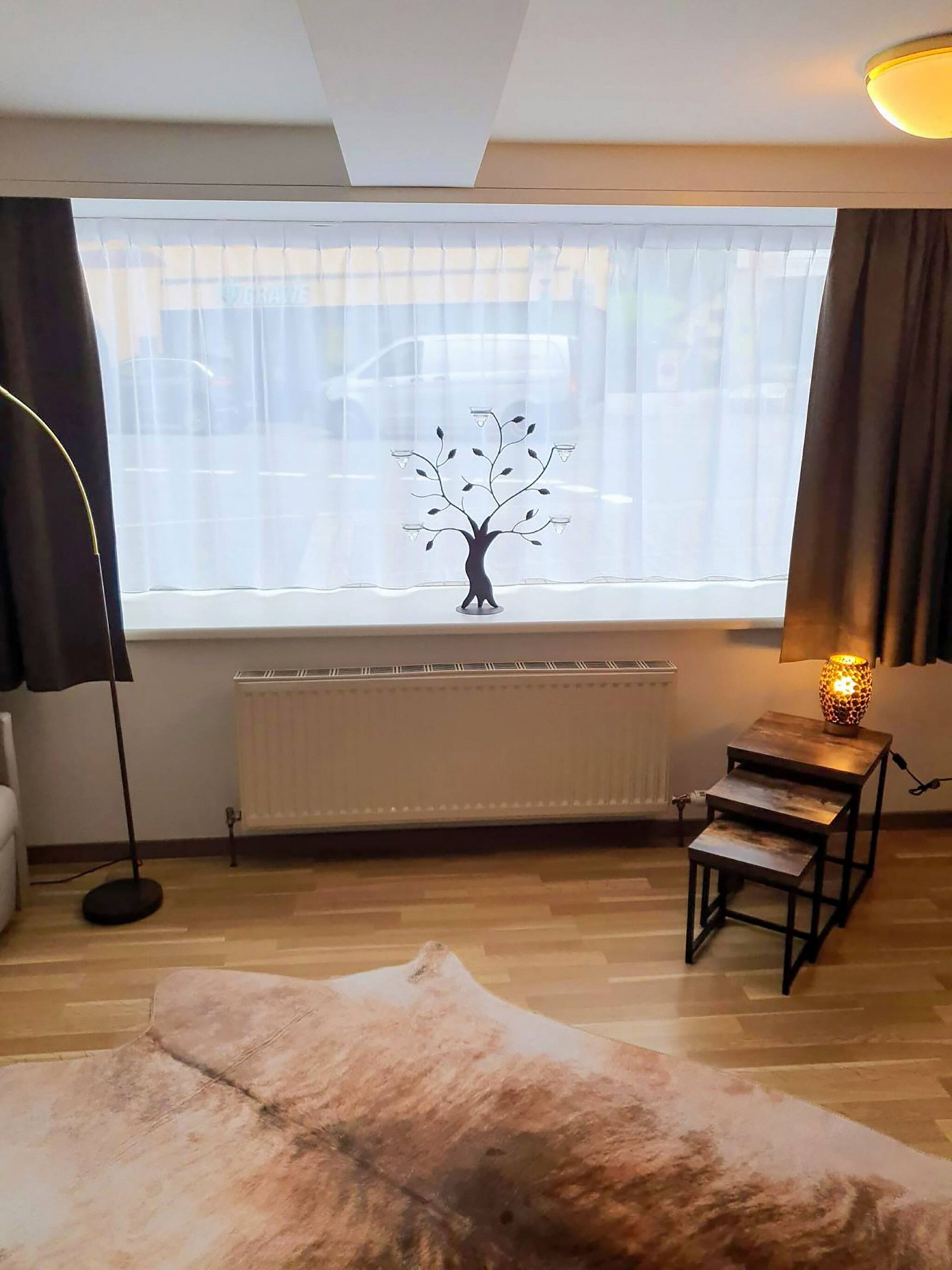 Ein Zimmer mit Heizkörper, Lampe auf einem kleinen Tisch, großem Fenster mit durchscheinenden Vorhängen und Stehlampe.