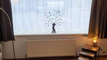 Ein Zimmer mit Heizkörper, Lampe auf einem kleinen Tisch, großem Fenster mit durchscheinenden Vorhängen und Stehlampe.