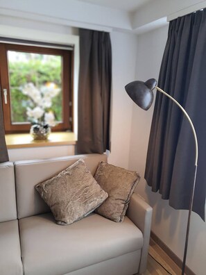 Ein modernes Wohnzimmer mit einer Couch, zwei Kissen, einer Stehlampe, einem Fenster mit Vorhängen und einer Vase mit Blumen.