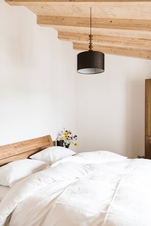 Un dormitorio con cabecera de madera, cama con ropa de cama blanca, lámpara colgante y un florero con flores sobre una mesita de noche.