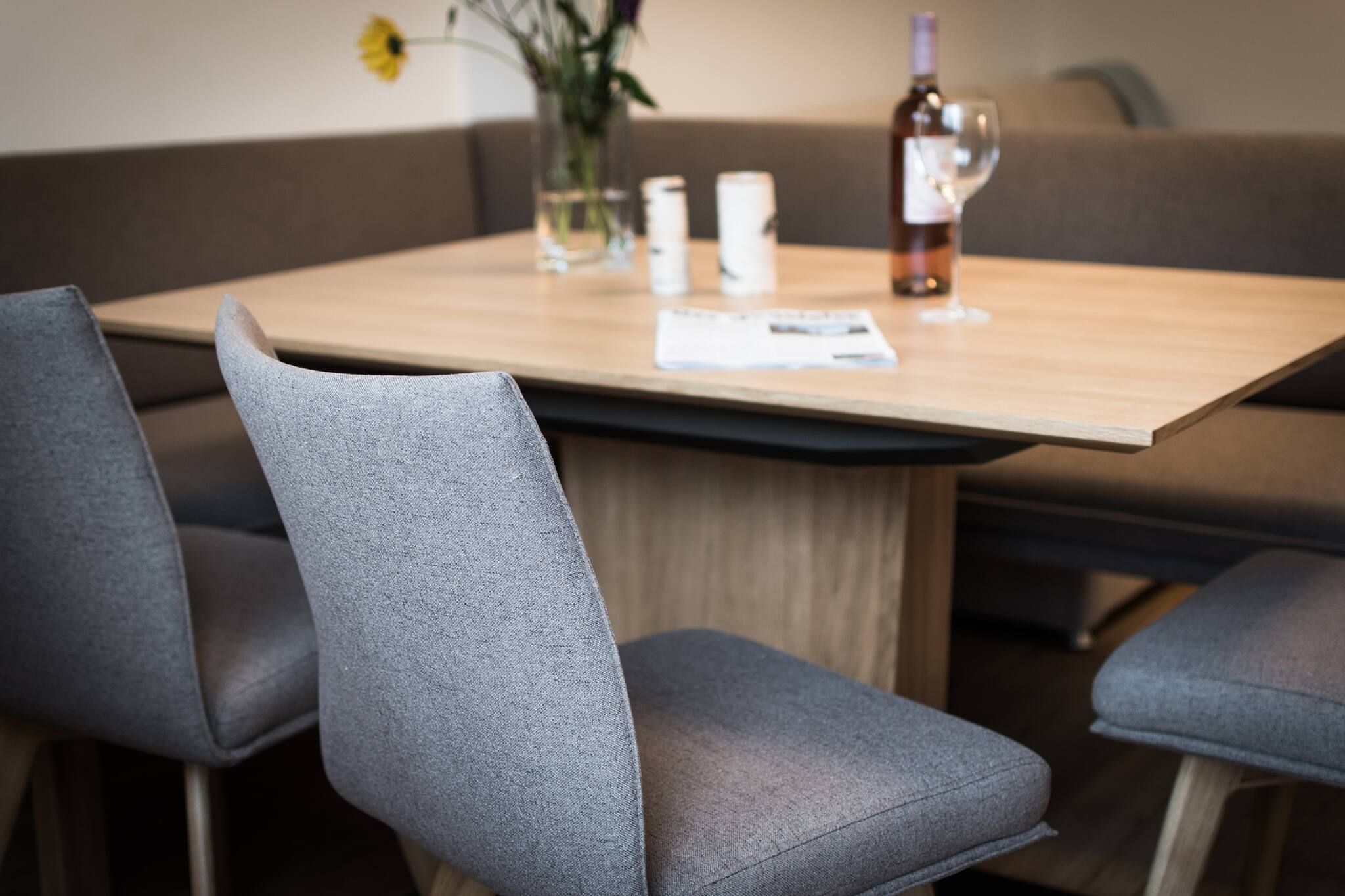 Un coin repas avec une table en bois, des chaises à revêtement gris, un vase contenant une fleur jaune et une bouteille de vin.